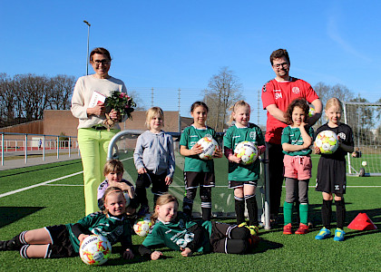 Sponsorenbild mit einer Gruppe junger Fußballerinnen, die neue Fußbälle in der Hand halten und gemeinsam mit ihrem Trainer ein Geschenk an die Sponsorin überreichen.