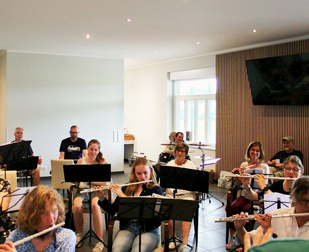 Gruppe an Musikerinnen, die gemeinsam musizieren, alle sitzen mit Notenständern und ihren Musikinstrumenten, viele spielen Querflöte