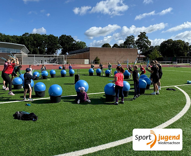 Beim polysportiven Camp werden große Pezzibälle als Trommeln benutzt, Alle Kinder stehen auf dem Sportplatz im Kreis, vor ihnen die Bälle, auf denen sie mit Stöcken trommeln