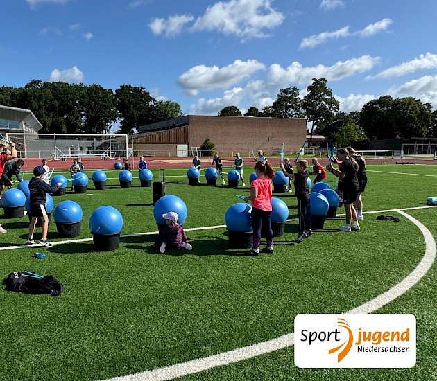 Beim polysportiven Camp werden große Pezzibälle als Trommeln benutzt, Alle Kinder stehen auf dem Sportplatz im Kreis, vor ihnen die Bälle, auf denen sie mit Stöcken trommeln