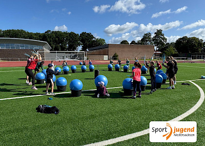 Beim polysportiven Camp werden große Pezzibälle als Trommeln benutzt, Alle Kinder stehen auf dem Sportplatz im Kreis, vor ihnen die Bälle, auf denen sie mit Stöcken trommeln