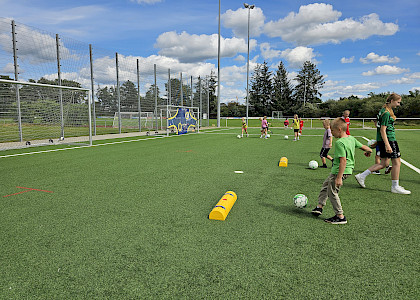 Kinder, die beim polysportiven Camp bei Sonnenschein Fußball spielen auf dem Kunstrasen