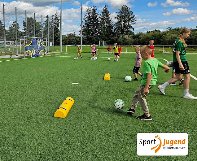 Kinder, die beim polysportiven Camp bei Sonnenschein Fußball spielen auf dem Kunstrasen