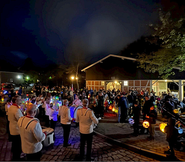 Übersicht über den Platz vorm Heimathaus mit viel Menschen, Kindern mit Laternen und dem spielenden Spielmannszug. Die Feuerwehr ist im Hintergrund