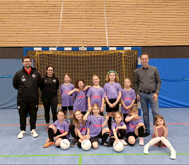Die F-Mädchen sitzen zusammen und der Sponsor und der Trainer stehen links und rechts neben ihnen. sie freuen sich über die neuen Trikots Die F-Mädchen sitzen zusammen und der Sponsor und der Trainer stehen links und rechts neben ihnen. sie freuen sich über die neuen Trikots