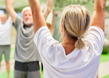 Eine Frau steht mit dem Rücken zum Fotografen und hebt die Arme beim Sport, im Hintergrund eine kleine Gruppe, die das nachmacht