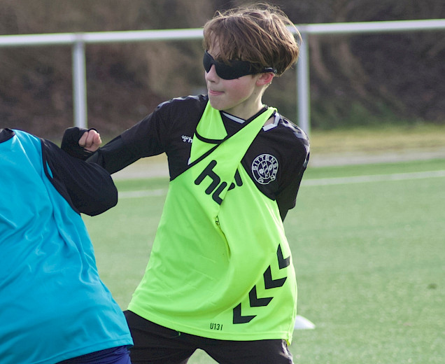 Auf dem Fußballplatz spielen 2 Jungs Fussball, beide tragen Leibchen und eine Verdunklungsbrille aus dem neurozentrierten Training