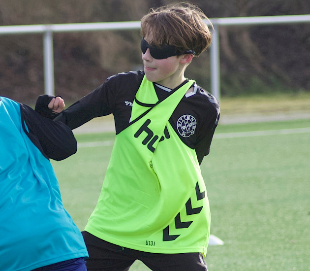 Auf dem Fußballplatz spielen 2 Jungs Fussball, beide tragen Leibchen und eine Verdunklungsbrille aus dem neurozentrierten Training Auf dem Fußballplatz spielen 2 Jungs Fussball, beide tragen Leibchen und eine Verdunklungsbrille aus dem neurozentrierten Training